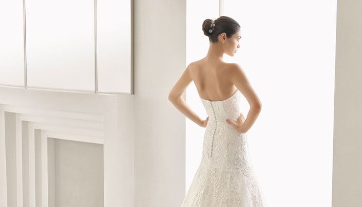 Rückenansicht einer Braut in einem schulterfreien Spitzenkleid, stehend vor einem hellen Fenster im Brauthaus Mannheim Heidelberg.