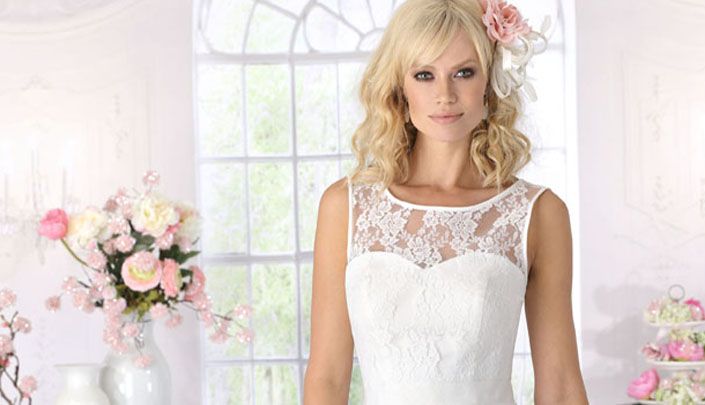 Braut in weißem Spitzenkleid mit zarter Blumenapplikation im Haar, stehend in einem hellen, blumengeschmückten Raum des Brauthauses Mannheim in Heidelberg.