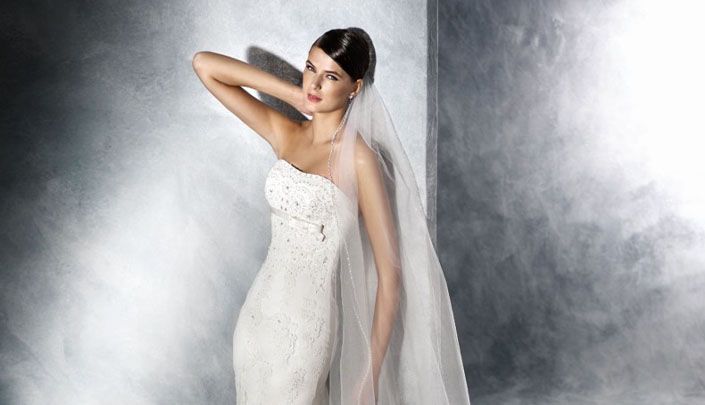 Braut in figurbetontem, trägerlosem Hochzeitskleid mit feinem Spitzenmuster und langem Schleier vor grauer Studio-Kulisse im Brauthaus Mannheim Heidelberg.